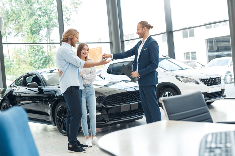 Young couple standing taking keys from new car from agent at salon smiling happy making deal